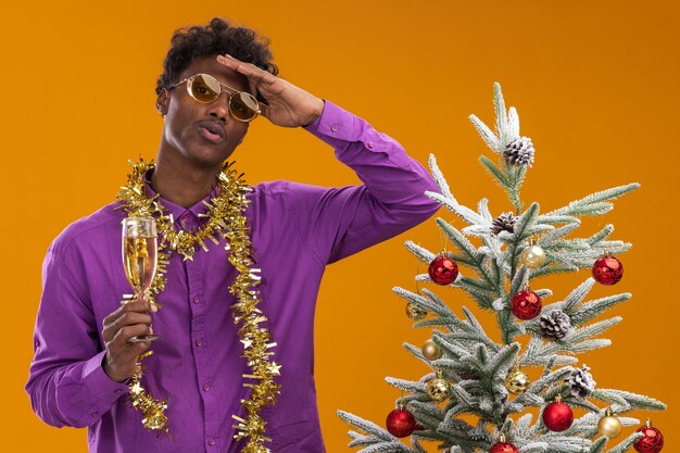 Impressed young afro-american man wearing glasses with tinsel garland around neck standing near decorated christmas tree holding glass of champagne 