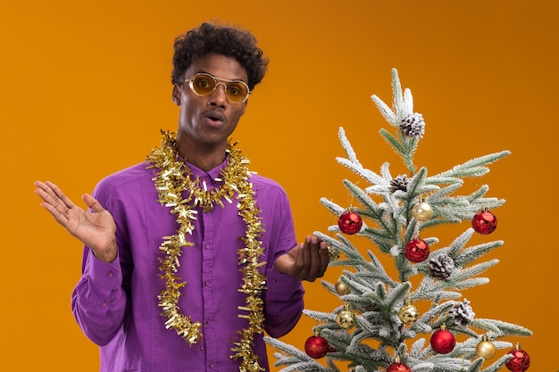 Impressed young afro-american man wearing glasses with tinsel garland around neck standing near decorated christmas tree on orange background