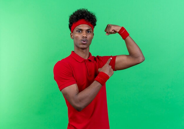 Impressed young afro-american sporty man wearing headband and wristband doing strong gesture and points isolated on green background