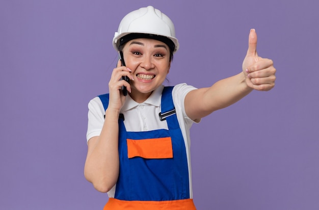 Впечатленная молодая азиатская девушка-строитель с белым защитным шлемом, разговаривающим по телефону и пальцем вверх