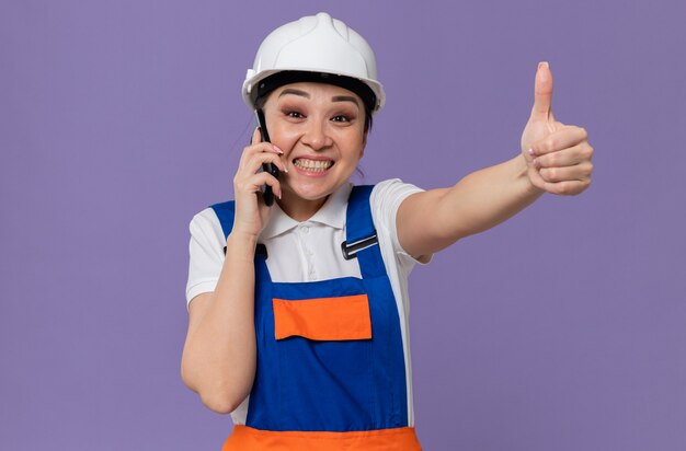 電話で話し、親指を立てる白い安全ヘルメットを持つ感銘を受けた若いアジアのビルダーの女の子