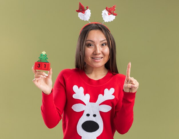 Impressed young asian girl wearing christmas hair hoop holding christmas toy points at up isolated on olive green background