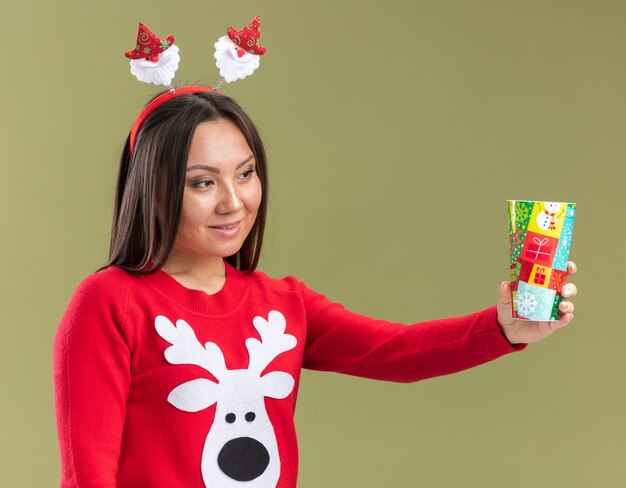 Free photo impressed young asian girl wearing christmas hair hoop holding out christmas cup at side isolated on olive green wall