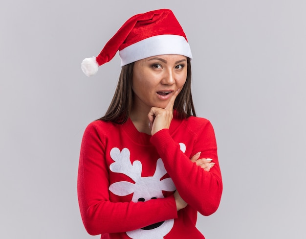 Impressed young asian girl wearing christmas hat with sweater putting finger on cheek isolated on white background