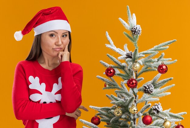 Impressed young asian girl wearing christmas hat with sweater standing nearby christmas tree putting finger on cheek isolated on orange background