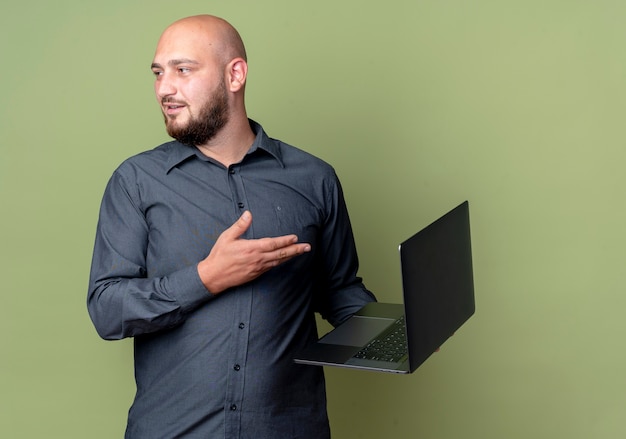 Бесплатное фото Впечатленный молодой лысый человек из колл-центра, держащий и указывающий рукой на ноутбук и смотрящий в сторону, изолированную на оливково-зеленом цвете с копией пространства