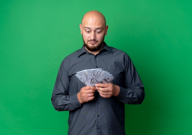 Impressed young bald call center man holding and looking at money isolated on green  with copy space