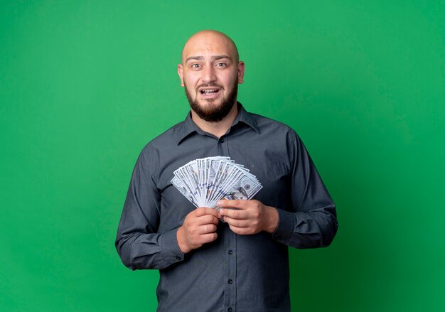 Impressed young bald call center man holding money isolated on green  with copy space