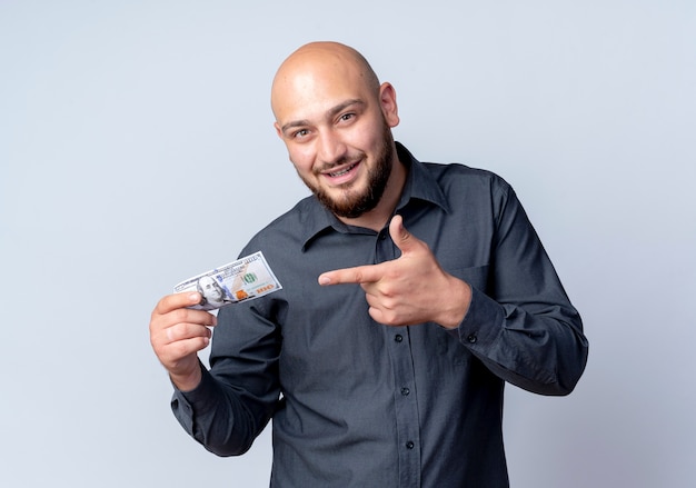 Impressed young bald call center man holding and pointing at money isolated on white 