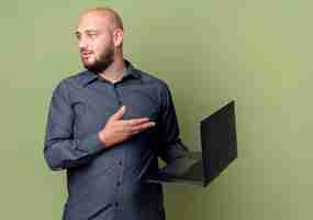 Free photo impressed young bald call center man holding and pointing with hand at laptop and looking at side isolated on olive green  with copy space