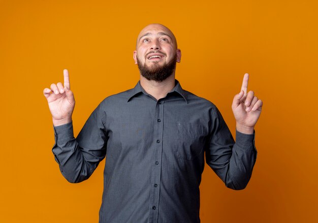 Impressed young bald call center man looking and pointing up isolated on orange 