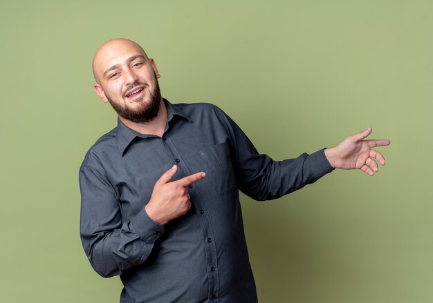 Impressed young bald call center man pointing at side isolated on olive green 