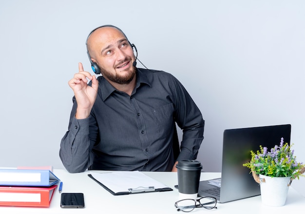 Impressed young bald call center man wearing headset sitting at desk with work tools looking and pointing up isolated on white 