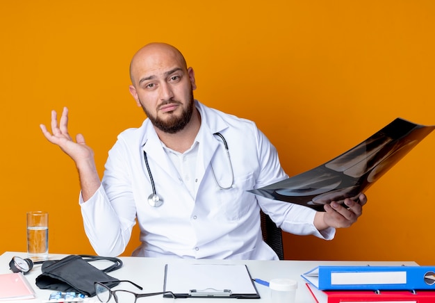 Impressionato giovane medico maschio calvo che indossa abito medico e stetoscopio seduto alla scrivania