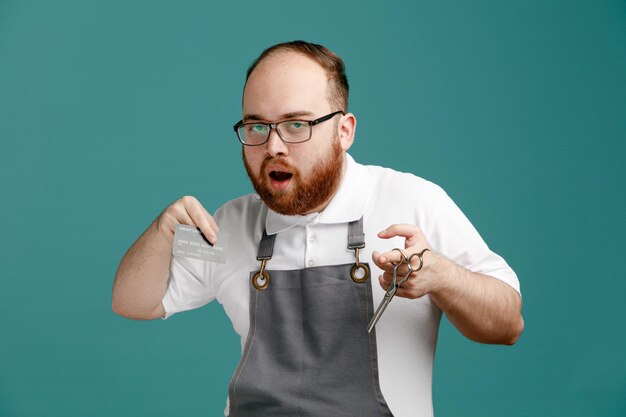 Impressed young barber wearing uniform and glasses holding scissors showing credit card looking at camera isolated on blue background