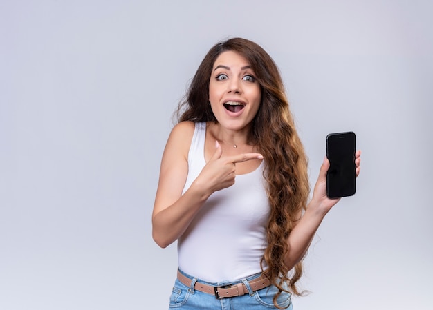 Impressed young beautiful girl holding mobile phone and pointing at it  with copy space