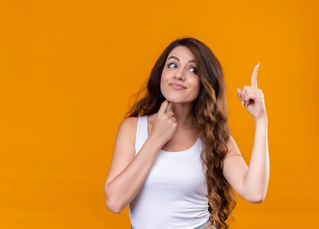 Impressed young beautiful girl pointing up putting finger under chin looking at right side on isolated orange space with copy space