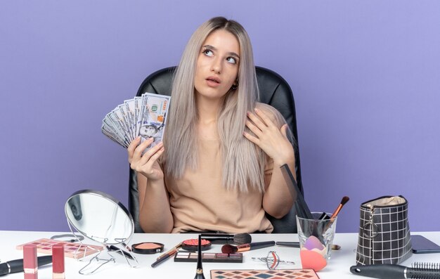 感銘を受けた若い美しい少女は、青い壁に分離された現金を保持している化粧ツールでテーブルに座っています。