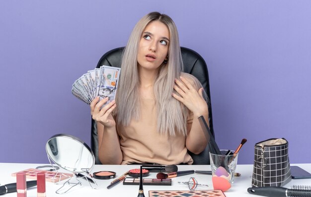 Impressed young beautiful girl sits at table with makeup tools holding cash isolated on blue wall