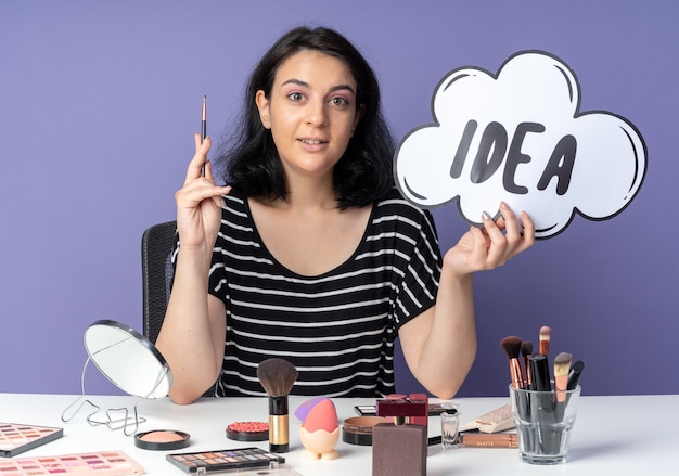 Impressed young beautiful girl sits at table with makeup tools holding idea bubble with makeup brush isolated on blue wall
