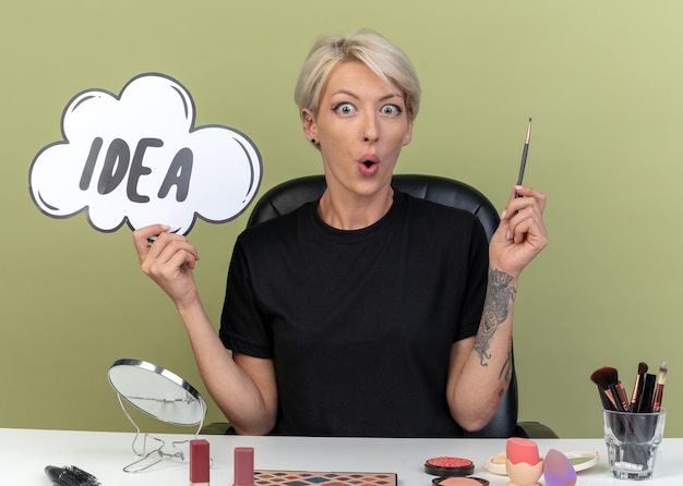 Free photo impressed young beautiful girl sits at table with makeup tools holding idea bubble with makeup brush isolated on olive green wall