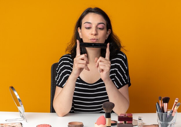 Впечатленная молодая красивая девушка сидит за столом с инструментами для макияжа, держа и глядя на кисть для пудры, изолированную на оранжевой стене