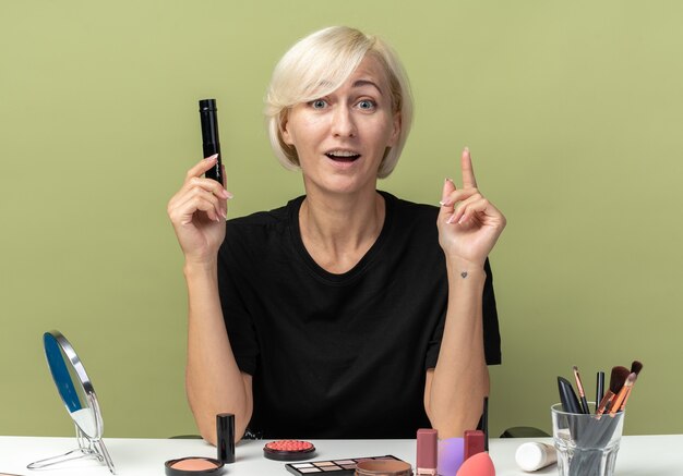 Impressed young beautiful girl sits at table with makeup tools holding mascara points at up isolated on olive green wall