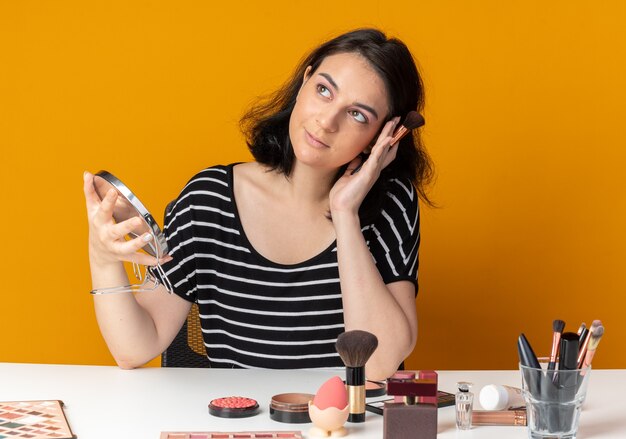 Impressed young beautiful girl sits at table with makeup tools holding mirror with powder brush isolated on orange wall