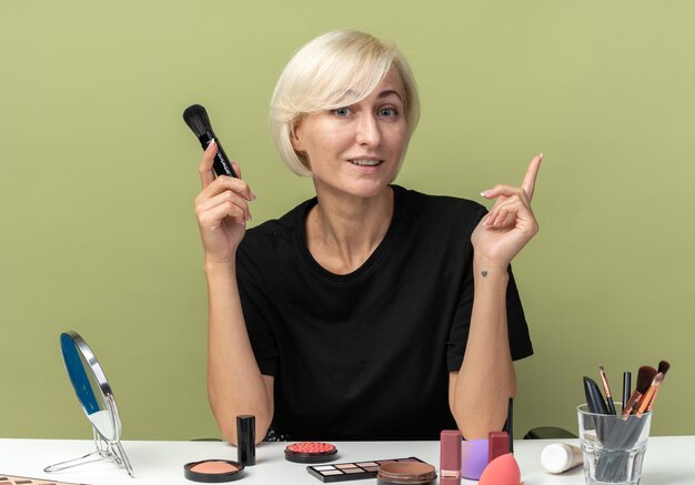 Impressed young beautiful girl sits at table with makeup tools holding powder brush points at up isolated on olive green wall