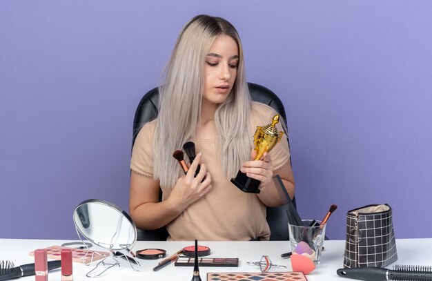 Impressed young beautiful girl sits at table with makeup tools holding winner cup with makeup brush isolated on blue wall