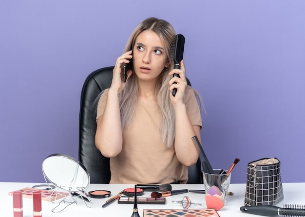 Впечатленная молодая красивая девушка сидит за столом с инструментами для макияжа, разговаривает по телефону, расчесывая волосы, изолированные на синей стене