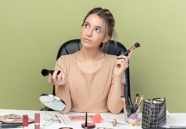 Впечатленная молодая красивая девушка, сидящая за столом с инструментами для макияжа, держа кисть для макияжа, изолированную на оливково-зеленой стене
