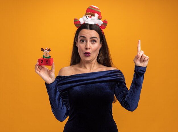 Impressed young beautiful girl wearing blue dress and christmas hair hoop holding toy points at up isolated on orange background