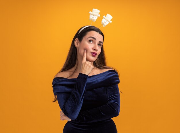 Impressed young beautiful girl wearing blue dress and christmas hair hoop putting finger on cheek isolated on orange background