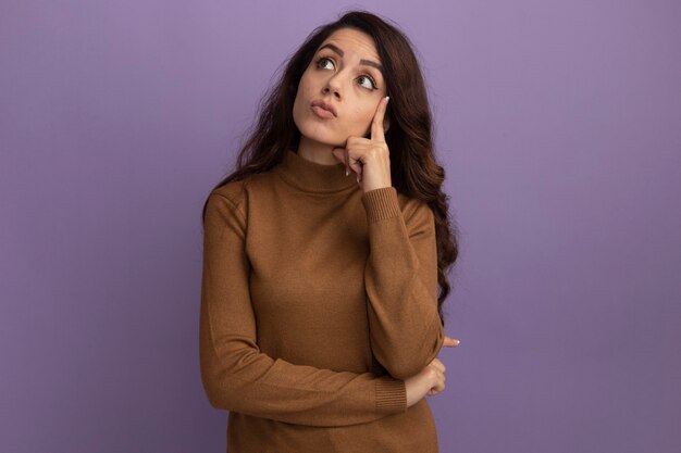 Впечатленная молодая красивая девушка в коричневом свитере с высоким воротом, положив палец на щеку, изолированную на фиолетовой стене