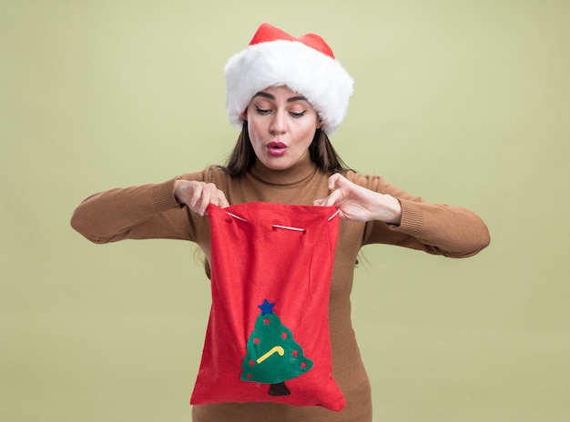 Impressed young beautiful girl wearing christmas hat holding and looking at christmas bag isolated on olive green background