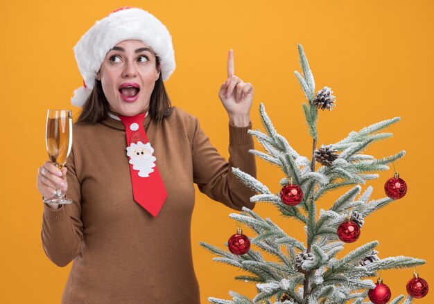 Impressed young beautiful girl wearing christmas hat with tie standing nearby christmas tree holding glass of champagne points at up isolated on orange background