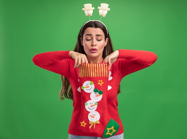 Foto gratuita impressionato giovane bella ragazza che indossa un maglione di natale con il cerchio di capelli di natale che tiene ed esamina i calzini di natale isolati sulla parete verde