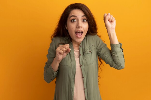 Impressed young beautiful girl wearing olive green t-shirt holding fists isolated on yellow wall