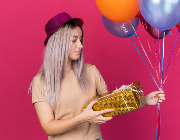 Impressed young beautiful girl wearing party hat holding balloons looking at gift box in her hand isolated on pink wall