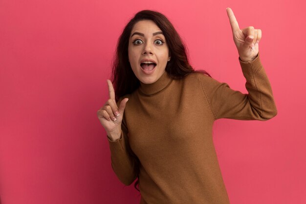 Impressed young beautiful girl wearing turtleneck sweater points at up isolated on pink wall