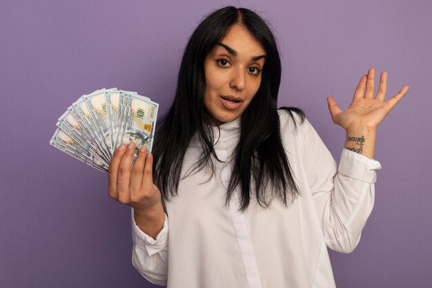 Free photo impressed young beautiful girl wearing white t-shirt holding cash spreading hand isolated on purple