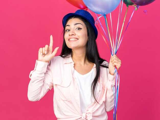 Impressed young beautiful woman wearing party hat holding balloons points at up isolated on pink wall