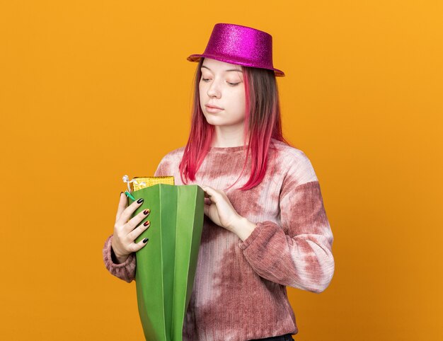 Impressed young beautiful woman wearing party hat holding and looking at gift bag isolated on orange wall