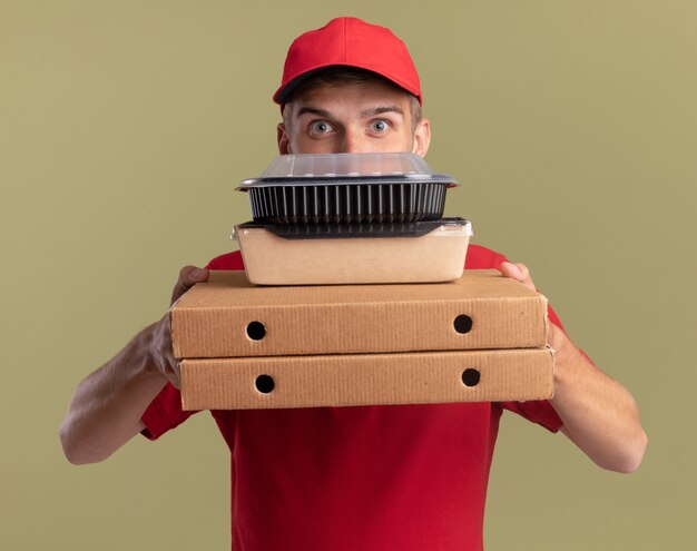 Impressed young blonde delivery boy holds food packages on pizza boxes isolated on olive green wall with copy space