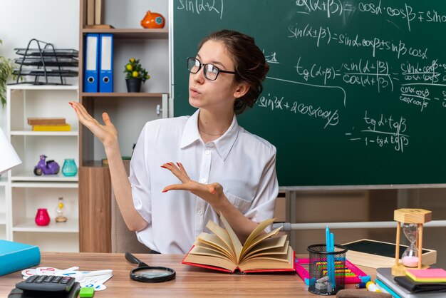 Impressed young blonde female math teacher wearing glasses sitting at desk with school tools looking and pointing with hands at side in classroom