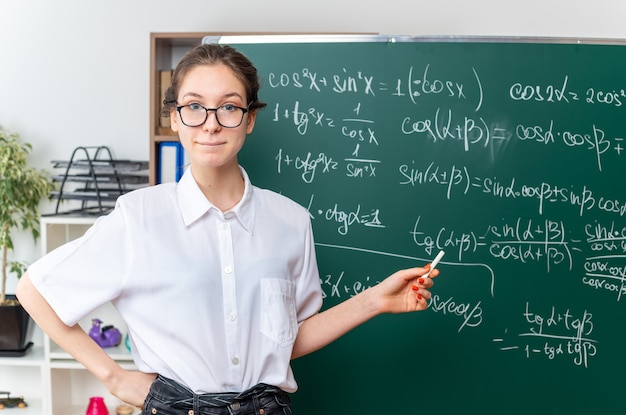 허리에 손을 유지하는 칠판에 쓰여진 수학 문제에 분필로 가리키는 카메라를보고 칠판 앞에 서있는 안경을 쓴 젊은 금발의 여성 수학 교사 감동