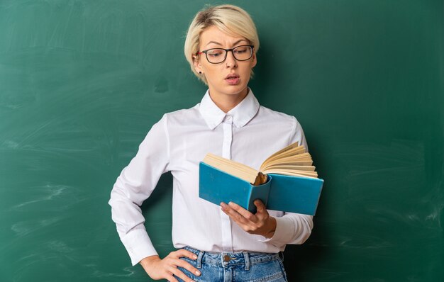 впечатленная молодая блондинка учительница в очках в классе, стоя перед классной доской, держа и читая книгу, держа руку на талии