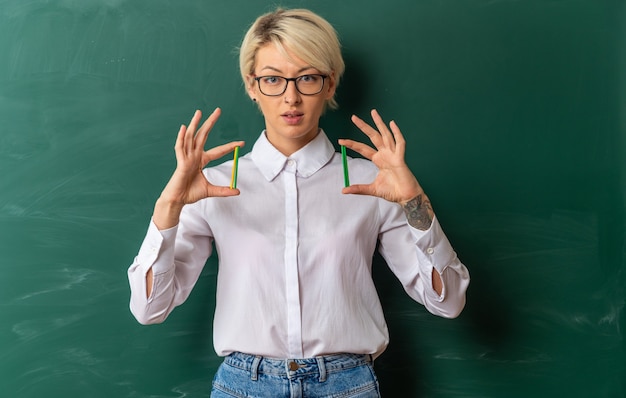 Впечатленная молодая блондинка учительница в очках в классе, стоя перед классной доской, показывая счетные палочки, глядя в камеру