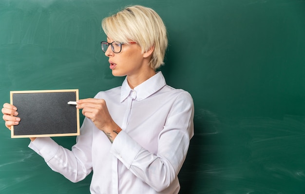 Impressed young blonde female teacher wearing glasses in classroom standing in front of chalkboard showing mini blackboard looking at it pointing at it with chalk with copy space